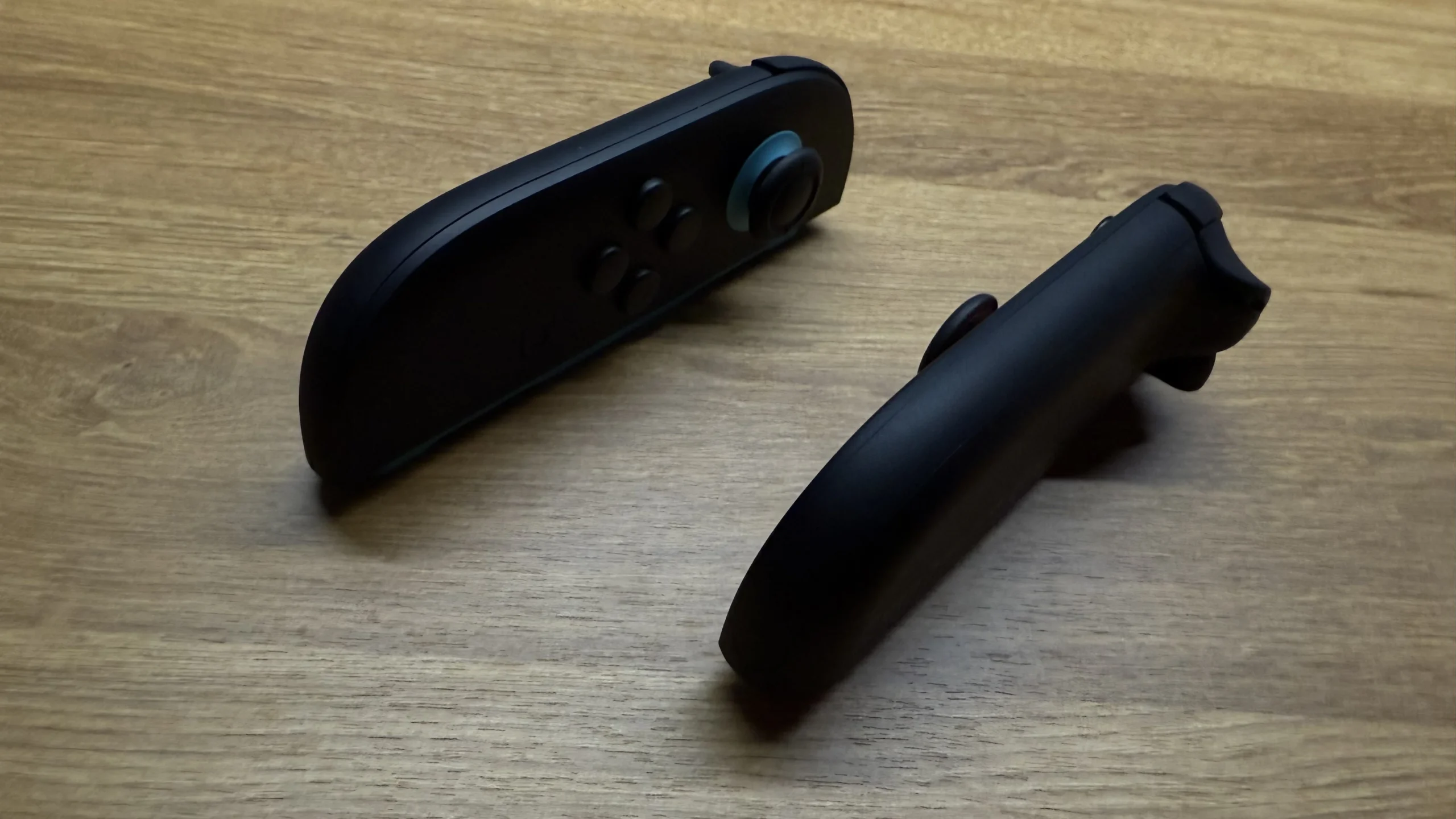 A pair of black Nintendo Switch 2 Joy-Cons resting on a wooden table. The left Joy-Con, with blue accents around its joystick, is angled to show its buttons and thumbstick, while the right Joy-Con is positioned sideways, highlighting its ergonomic curve. This setup depicts the "mouse mode" functionality.