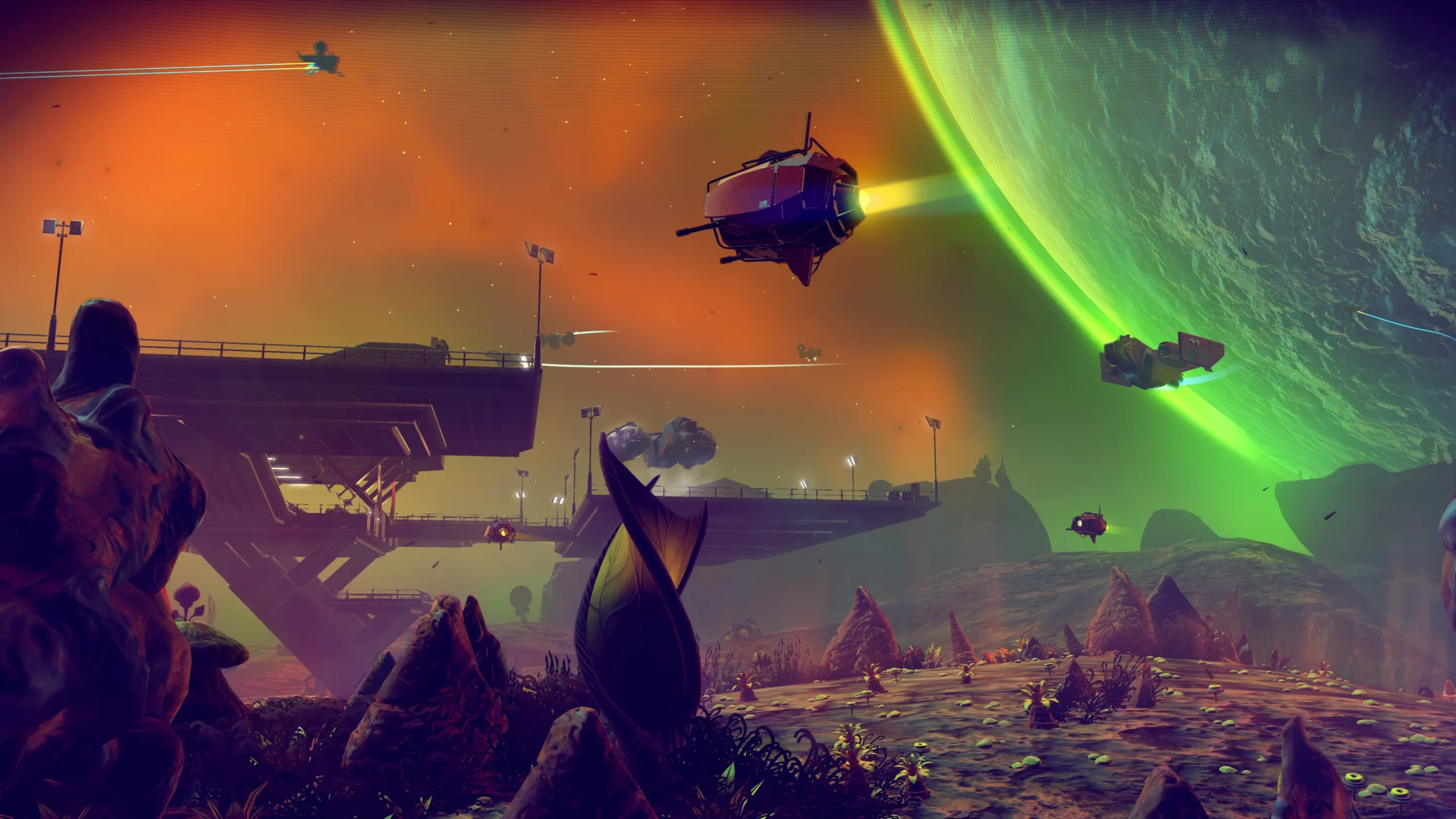 A wide-angle, stylized screenshot from No Man's Sky depicting an alien planet at twilight. The foreground features unusual rock formations and sparse alien flora. In the mid-ground, a massive, dark structure spans across the landscape. Several unique spacecraft are seen flying through the vibrant orange and purple sky, with a large, green glowing planet dominating the background.
