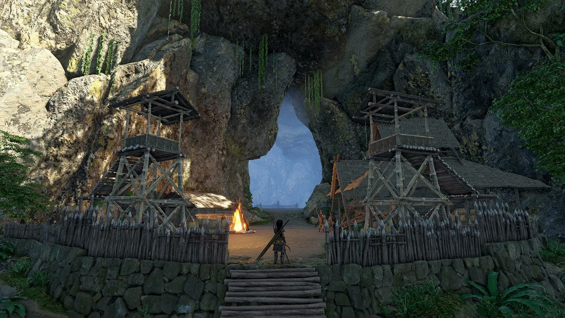 A tribal settlement with wooden watchtowers and a campfire, built into the base of a large rocky cliff face with a glowing cave entrance in the background, from the game Soulmask.